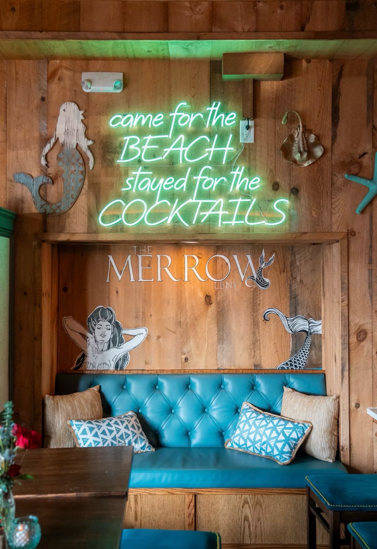 The Merrow's back banquette. Cyan neon reading 'came for the BEACH stayed for the COCKTAILS' over a teal tufted booth.